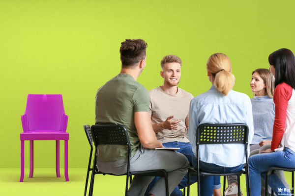 Eine im Bild angeschnittene Gruppe im Kreis sitzender Menschen unterhält sich, während ein einzelner, leerer Stuhl abseits steht.