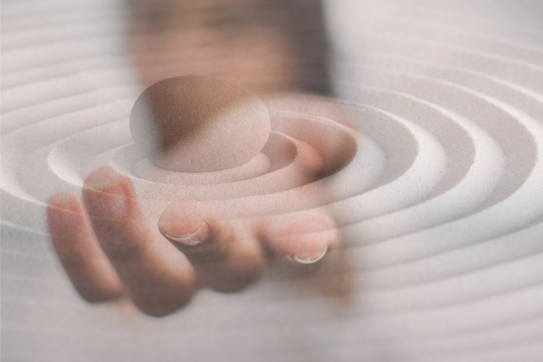 Eine Hand hält einen glatten Stein über konzentrischen Kreisen im Sand alsSymbol für Achtsamkeit und innere Ruhe