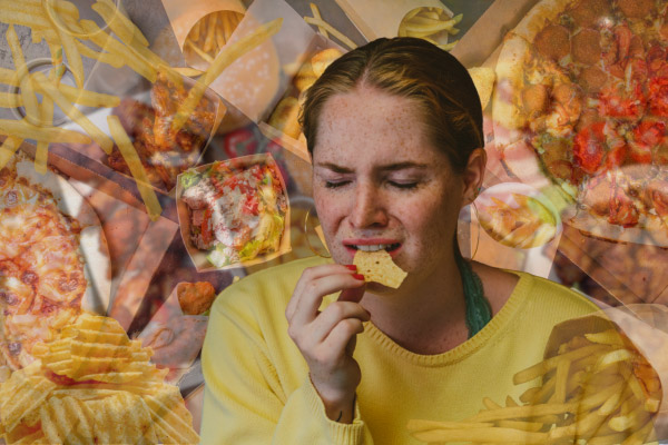 Eine junge Frau isst Chips und schaut verzweifelt, im Hintergrund ist eine große Menge ungesunder Lebensmitteln gestapelt