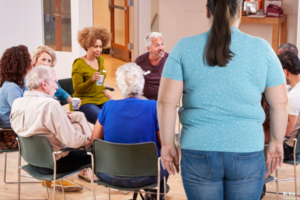 Übergewichtige Frau betritt einen Stuhlkreis einer therapeutischen Selbsthilfegruppe und blickt auf die bereits miteinander sprechenden Teilnehmenden