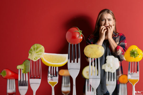 Frau mit panischem Gesichtsausdruck steht vor einer Reihe von Gabeln, auf denen verschiedene gesunde Lebensmittel wie Gemüse und Obst stecken. Die Szene symbolisiert Angst und Stress bei Orthorexie.