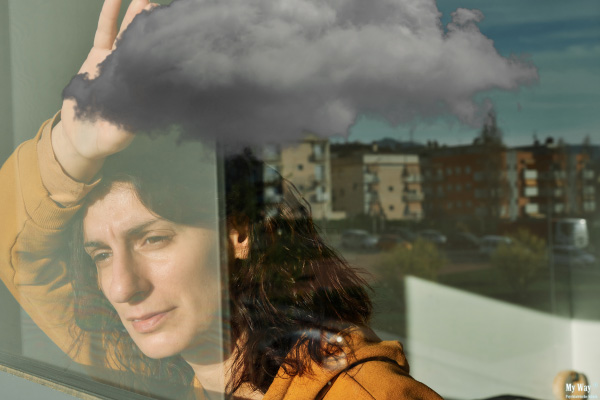 Nachdenkliche Frau blickt aus dem Fenster, ihr Spiegelbild zeigt eine dunkle Gewitterwolke über dem Kopf als Symbol für psychische Belastung