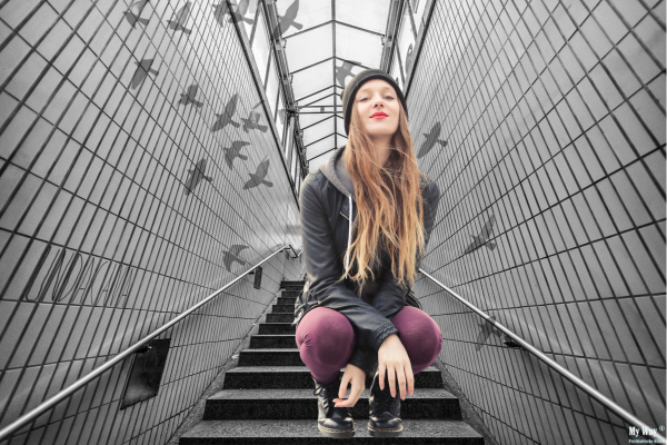 Junge Frau mit langem Haaren und Mütze hockt auf einer Treppe in einem modernen, gekachelten Tunnel. Im Hintergrund fliegen stilisierte Vögel, was eine surreale Atmosphäre schafft